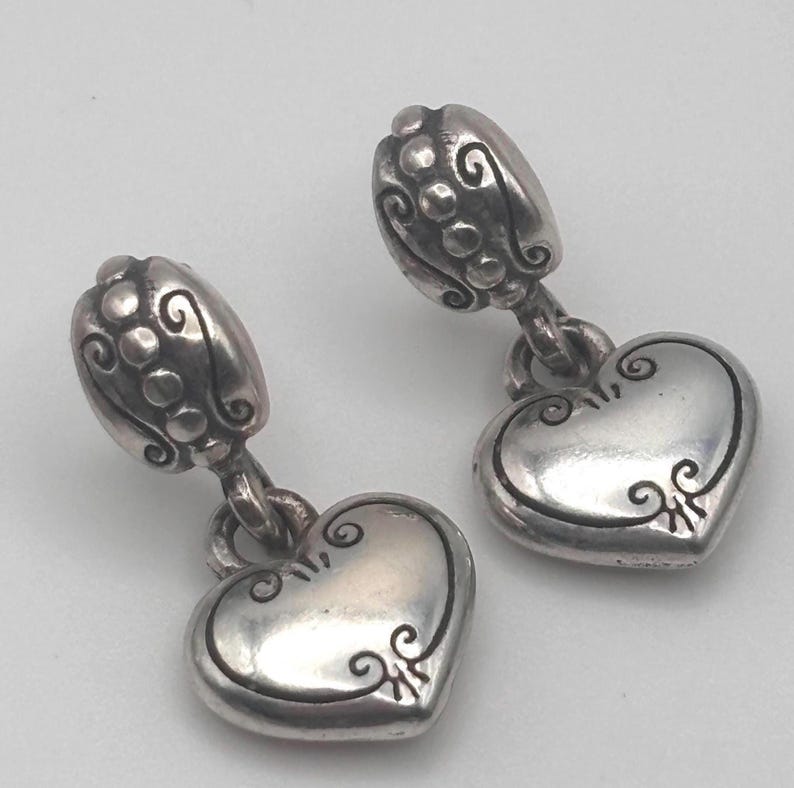 Op de afbeelding: Een paar zilverkleurige oorhangers. Elke oorbel heeft een hartvormige bedel met decoratieve krullen en een bloemvormige bovenkant. De oorbellen hebben een vintage design.