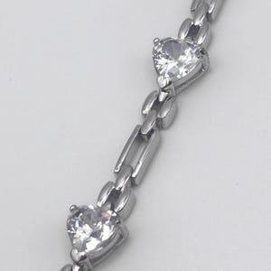 May include: A silver bracelet with a link chain design. The bracelet features two heart-shaped, clear gemstones. The gemstones are set in a silver metal setting. The bracelet is displayed against a white background.
