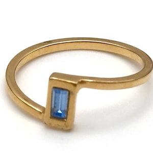 May include: A gold-colored ring with a rectangular, light blue gemstone. The ring has a unique, angular design, with the gemstone set in a rectangular bezel. The band is smooth and rounded.