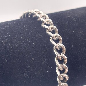 Puede incluir: Un brazalete plateado con un diseño de eslabones en forma de corazón. El brazalete de metal texturizado se muestra sobre un fondo oscuro. Los eslabones están entrelazados, creando una cadena.