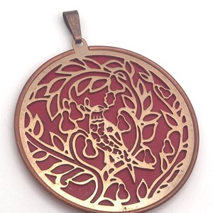 May include: A circular pendant featuring a metallic gold-toned cutout design of a bird perched among leafy branches against a solid deep red background. The pendant has a copper-colored outer rim and a matching metal bail at the top for hanging.