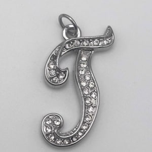 May include: Silver-toned pendant in the shape of the letter "T", encrusted with numerous small, clear crystals. The pendant has a loop at the top for a chain. The crystals are set in a curved, cursive design, creating a sparkling effect.