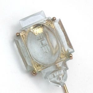 May include: A clear glass stick pin featuring a rectangular faceted design with a central oval inlay. The glass is set within a gold-toned metal frame, accented by small metallic spheres at the corners. A thin gold-toned metal pin extends from the base.