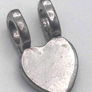 May include: A silver-colored heart-shaped pendant with two rounded loops at the top. The pendant has a slightly textured surface and a simple, classic design, suitable for jewelry making or as a charm.