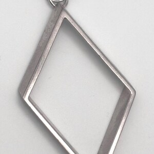 May include: A silver-toned metal pendant in the shape of a hollow diamond or rhombus. The frame has a smooth, flat surface and a consistent thickness throughout. A small, circular jump ring is attached to the top point for hanging purposes.