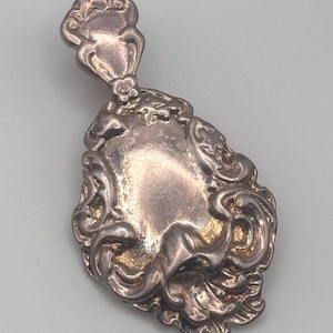 May include: Ornate silver pendant with a detailed, symmetrical design. The pendant features a central shield shape surrounded by scrolling embellishments and a decorative top element. The metal has a polished, antique finish.