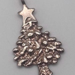May include: A silver-toned Christmas tree pendant with a star on top. The tree has a textured, bubbly design and a detailed trunk. The pendant is likely made of metal and is suitable for a necklace or charm bracelet.