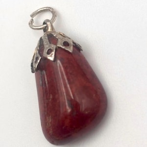 May include: A red gemstone pendant with a silver-colored metal cap and loop. The pendant is a teardrop shape and has a polished, glossy finish. The metal cap has a decorative design. The pendant is likely for a necklace or bracelet.