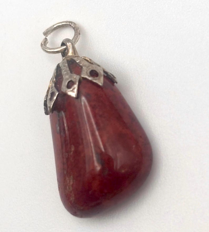 May include: A red gemstone pendant with a silver-colored metal cap and loop. The pendant is a teardrop shape and has a polished, glossy finish. The metal cap has a decorative design. The pendant is likely for a necklace or bracelet.