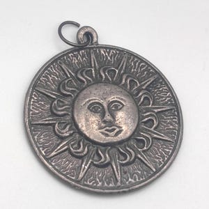 May include: A circular silver-toned metal pendant featuring a raised sun face at the center. The sun has stylized facial features and is surrounded by alternating straight and wavy rays. A small metal jump ring is attached to the top of the pendant.