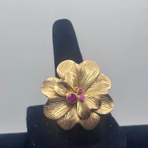 May include: A gold-toned ring featuring a large, textured flower design with three pink rhinestones in the center.