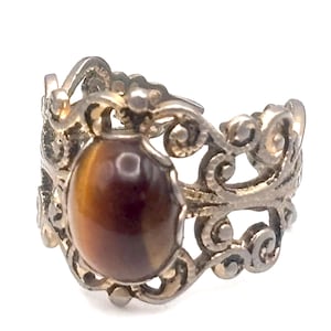 May include: A silver-toned ring with an oval, brown gemstone at the center. The ring features an ornate, openwork design with swirling patterns. The gemstone has a polished, reflective surface, and the ring is set against a white background.