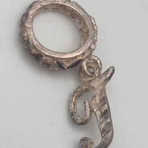 May include: A silver-colored charm with a ring and a cursive letter "J" pendant. The ring has a decorative border. The letter "J" has a textured, striped design. The charm is likely made of metal.