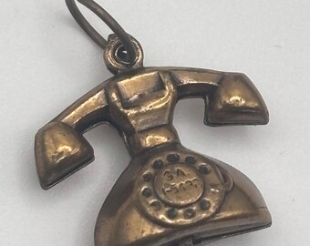 May include: A vintage bronze-colored telephone charm with a loop for attaching to a necklace or bracelet. The charm features a rotary dial and a classic handset design. The dial has the numbers 34 and 5433. The charm is a small, detailed piece of jewelry.