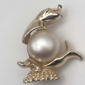 May include: A gold-toned vintage brooch in the shape of a seal balancing a large, pearlescent faux pearl. The seal is detailed with flipper-like arms and a textured base. The brooch has a classic, elegant design.