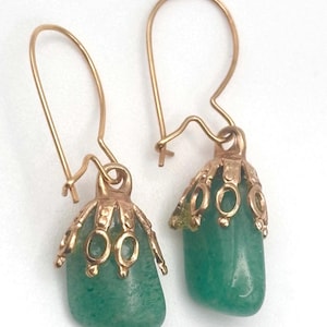 May include: A pair of gold-colored earrings with green gemstone pendants. The earrings feature a decorative gold cap with oval cutouts. The green gemstones are smooth and rounded. The earrings have a simple hook closure.