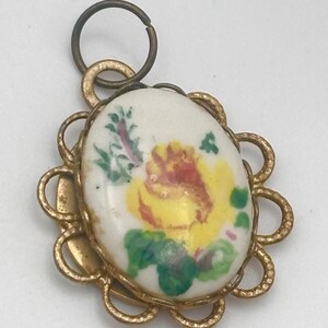 May include: Oval pendant with a hand-painted yellow rose on a white background, framed by a gold-colored scalloped edge. A small loop and ring are at the top for attachment. The pendant is likely a charm or jewelry component.