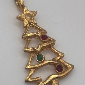 May include: Gold-toned Christmas tree pendant with a star on top and three colored gemstones. The pendant features a textured finish and a loop for a chain. The gemstones are red and green. A festive holiday jewelry item.