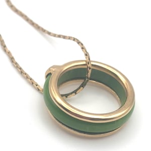 May include: A gold-toned necklace featuring a circular pendant with a green jade-like inlay. The pendant is suspended from a delicate chain. The necklace is set against a white background.