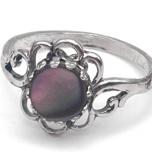 May include: A silver ring with a dark purple, iridescent center stone. The ring features a floral design around the stone and decorative flourishes on the band. The ring is set against a white background.