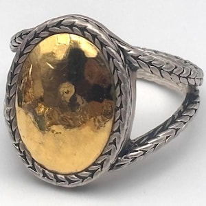 May include: Oval-shaped ring with a gold-colored center and a silver-colored braided band. The gold center has a hammered texture. The band has a braided design. The ring is set against a white background.