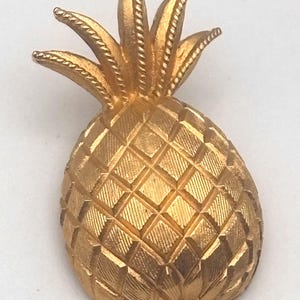 May include: A gold-colored pineapple-shaped brooch. The brooch has a textured surface with a diamond pattern on the body and detailed leaves at the top. The pineapple is a classic symbol of hospitality and tropical themes.