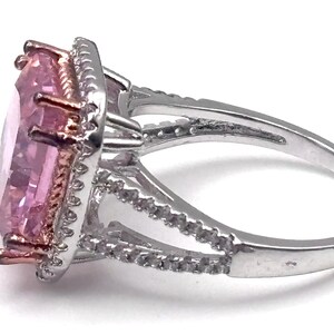 May include: A silver ring with a large, light pink, rectangular gemstone. The gemstone is surrounded by a halo of smaller, clear stones. The ring has a split shank design and is set against a white background.