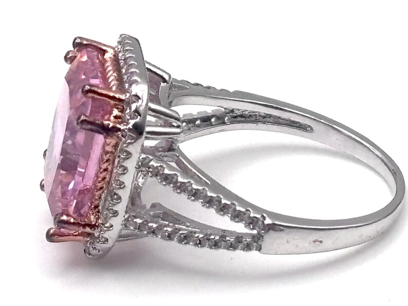 May include: A silver ring with a large, light pink, rectangular gemstone. The gemstone is surrounded by a halo of smaller, clear stones. The ring has a split shank design and is set against a white background.