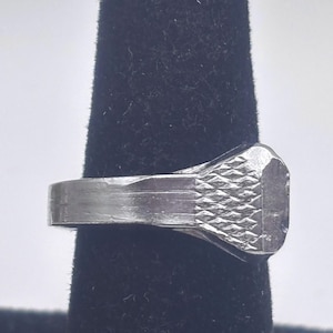 May include: A silver-colored ring with a rectangular face featuring a textured, diamond-patterned design. The ring is set against a black background, highlighting its metallic sheen and geometric details. The band is smooth and simple.