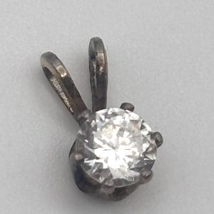 May include: A silver pendant featuring a large, round, clear gemstone. The pendant has a simple design with a loop for a chain and a setting that holds the gemstone securely. The gemstone sparkles brightly against the silver.