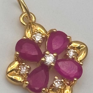 May include: A gold-colored pendant with a floral design. The pendant features five teardrop-shaped, deep red gemstones arranged around a central, round, clear gemstone. Small, clear gemstones are also set between the red gems.