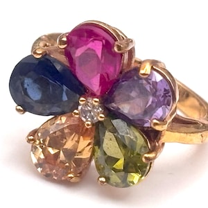 May include: A gold ring featuring a flower design with six colorful gemstones. The gemstones are pear-shaped and include pink, blue, purple, orange, green, and a small diamond in the center. The ring is set against a white background.