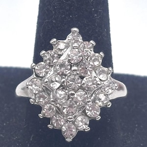 May include: A silver-toned ring featuring a cluster of clear, round rhinestones. The rhinestones are arranged in a floral or starburst pattern, creating a sparkling effect. The ring is set on a black velvet surface.