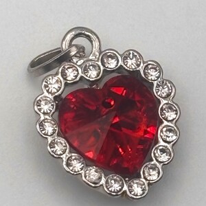 May include: A heart-shaped pendant with a large, faceted red crystal center. The crystal is surrounded by a silver-colored metal frame with a border of small, clear crystals. The pendant has a loop for attaching to a chain.