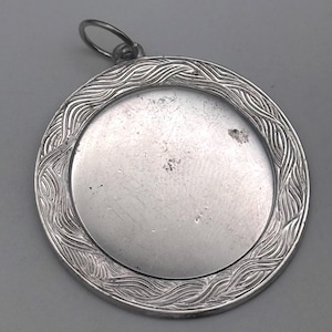May include: A round, silver-colored pendant with a textured, wave-like border. The pendant has a smooth, circular center and a small loop at the top for hanging. The metal has a slightly aged appearance.