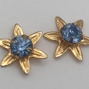 May include: Two gold-toned star-shaped earrings, each featuring a light blue gemstone center. The earrings have a detailed, textured design on the star points. The gemstones are round and faceted, reflecting light. The background is a plain, light color.