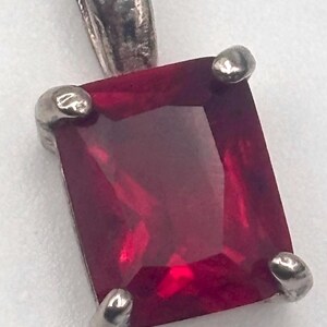 May include: A square-cut, deep red gemstone pendant with a silver bail. The gemstone is set in a silver bezel with four rounded prongs. The pendant is likely for a necklace or bracelet.