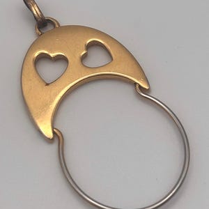 May include: A gold-colored pendant with a crescent-shaped top featuring two heart cutouts. A silver-colored circular loop is attached to the bottom of the pendant. The pendant has a small loop at the top for a chain.