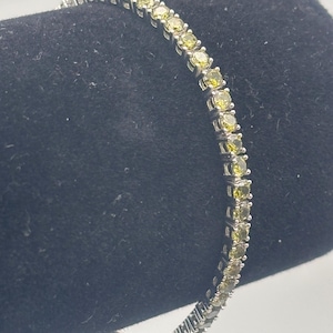 May include: A silver bracelet with a row of small, round, light green gemstones. The bracelet is displayed against a black background, showcasing the sparkle of the stones. The bracelet is a delicate piece of jewelry.