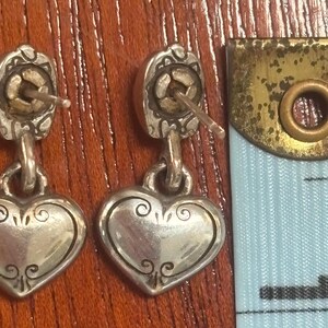 Op de afbeelding: Een paar zilverkleurige hartvormige oorhangers. Elke oorbel heeft een decoratieve bovenkant met een pin en een hartvormige hanger met krullen. Een meetlint staat op de achtergrond.