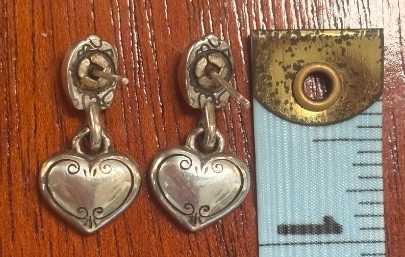 Op de afbeelding: Een paar zilverkleurige hartvormige oorhangers. Elke oorbel heeft een decoratieve bovenkant met een pin en een hartvormige hanger met krullen. Een meetlint staat op de achtergrond.