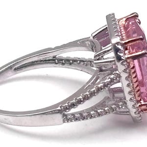 May include: A silver ring with a large, rectangular pink gemstone. The gemstone is surrounded by a halo of smaller, clear stones. The ring band features additional small stones. The setting is a mix of silver and rose gold.