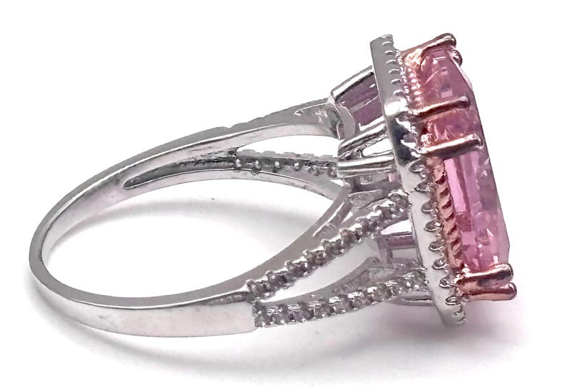 May include: A silver ring with a large, rectangular pink gemstone. The gemstone is surrounded by a halo of smaller, clear stones. The ring band features additional small stones. The setting is a mix of silver and rose gold.