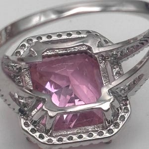 May include: A close-up of a silver ring featuring a large, rectangular pink gemstone. The gemstone is surrounded by a halo of smaller, clear stones. The ring's band is also silver.