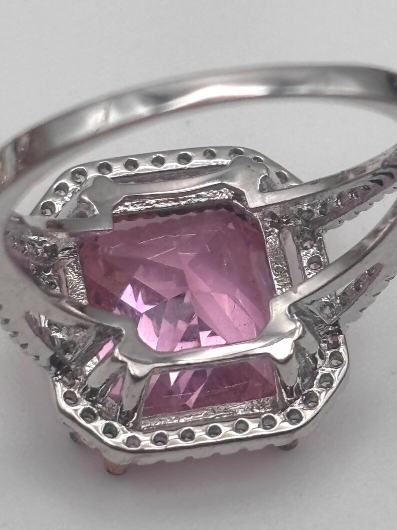 May include: A close-up of a silver ring featuring a large, rectangular pink gemstone. The gemstone is surrounded by a halo of smaller, clear stones. The ring's band is also silver.