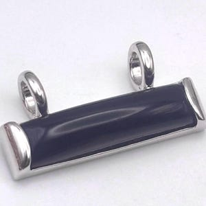 May include: A rectangular pendant featuring a smooth, polished black stone inlay set within a shiny silver-toned metal frame. The piece includes two vertical silver-toned loops at the top, designed for attachment to a chain or cord necklace.