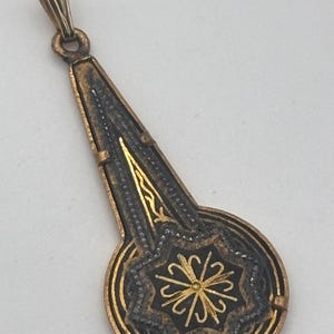 May include: A vintage pendant with a gold-colored bail and a long, ornate body. The pendant features a circular base with a starburst design and intricate detailing in gold and black. The overall design is symmetrical and elegant.