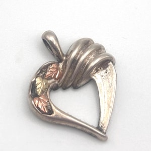 May include: Silver-tone heart pendant with a twisted design and three small leaf accents in gold and rose gold. The pendant has a loop for a chain and is set against a white background.