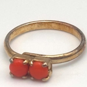 May include: A gold-toned metal ring featuring two small, round, vibrant orange stones set side-by-side in a prong setting. The band is thin and circular, showing a metallic finish with subtle signs of wear along the outer surface of the ring.