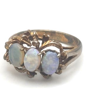 May include: A metal ring featuring three oval-shaped opals set in a decorative, filigree-style band. The opals display iridescent flashes of blue and white, while the metal band has a tarnished, aged appearance with intricate leaf-like details.